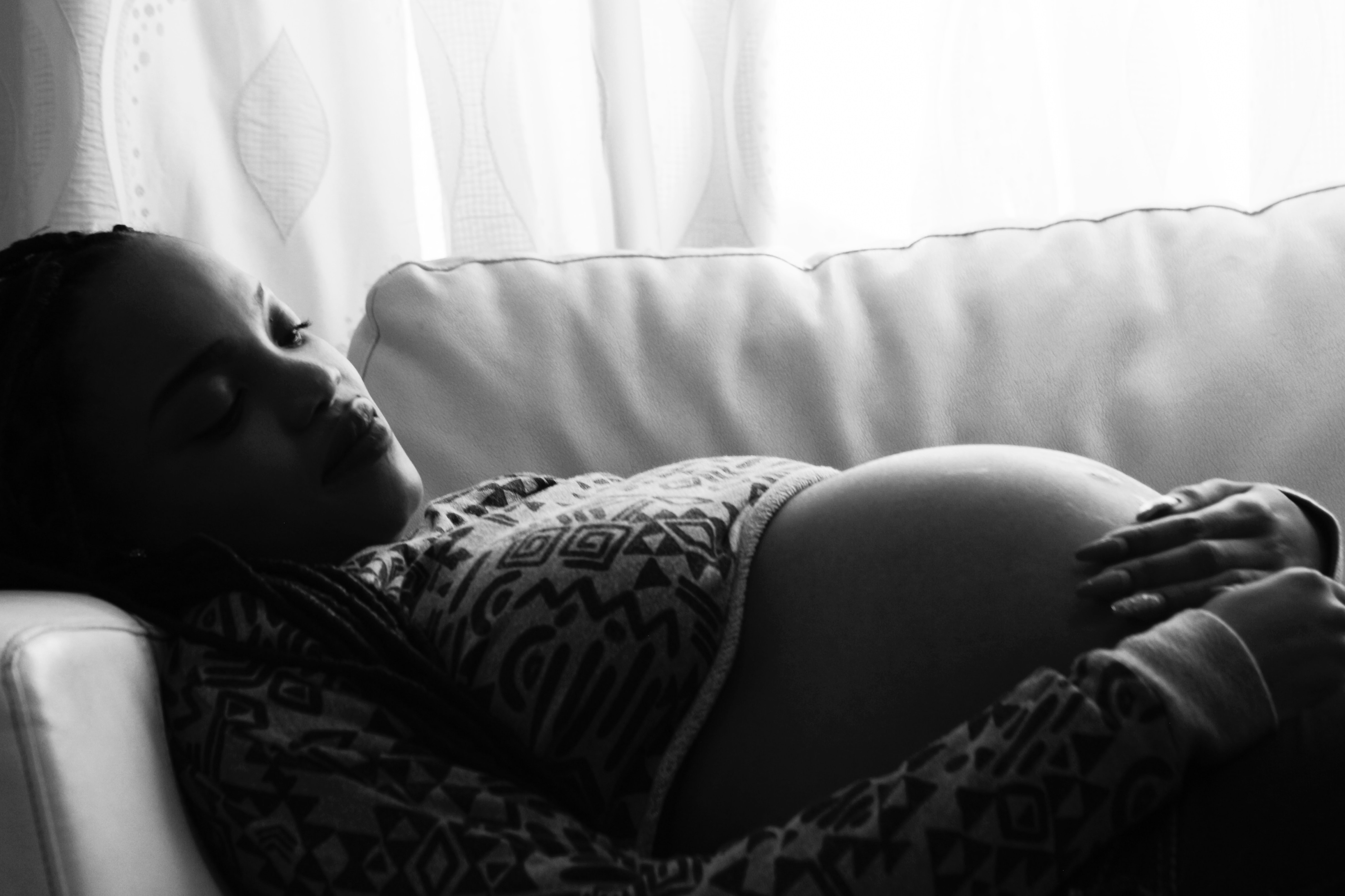 A Black pregnant woman recilines on a sofa by a window.